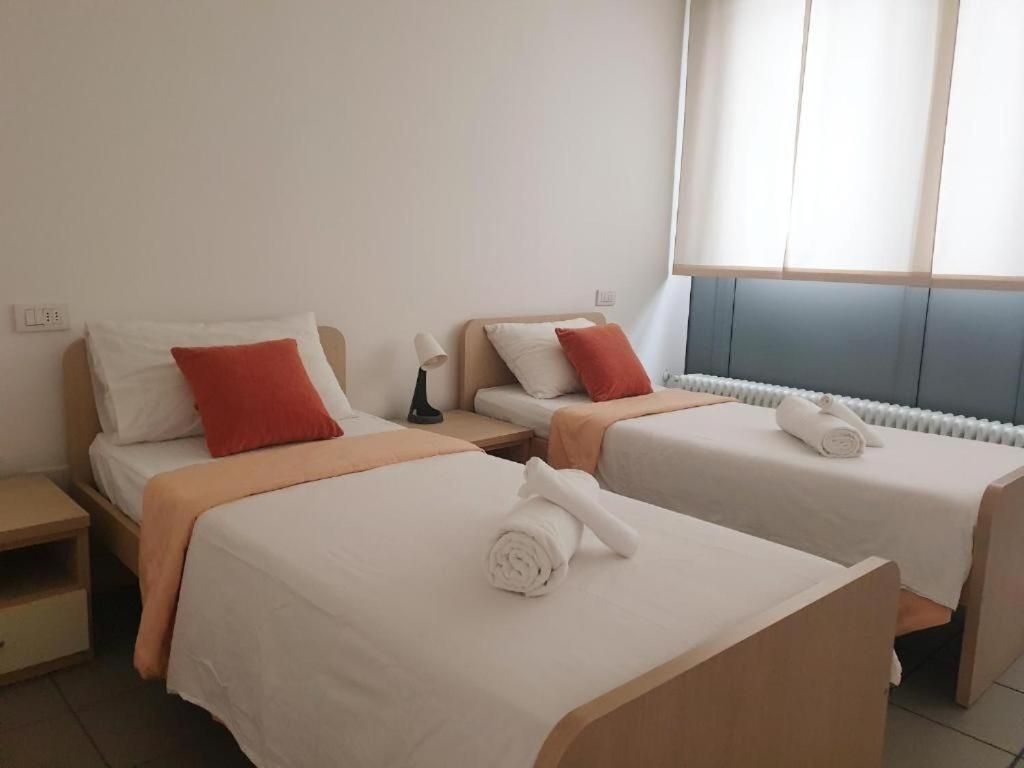 A hotel room with two beds, each with white bedding and red pillows, a bedside table with a lamp, and a window with blinds.