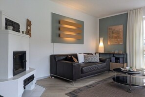 A modern living room with a fireplace, a black leather sofa, a side table, and a wall-mounted light fixture. - Holiday apartment with 1 bedroom (Füssen)