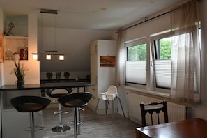 A modern kitchen with a central island, bar stools, a dining table, and a window with blinds. - Holiday apartment with 1 bedroom (Schneverdingen)