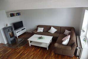 A living room with a brown sofa, white cushions, a white coffee table, a flat-screen TV on a white stand, and a small white cabinet above the TV. - Holiday home with 3 bedrooms (Greetsiel)