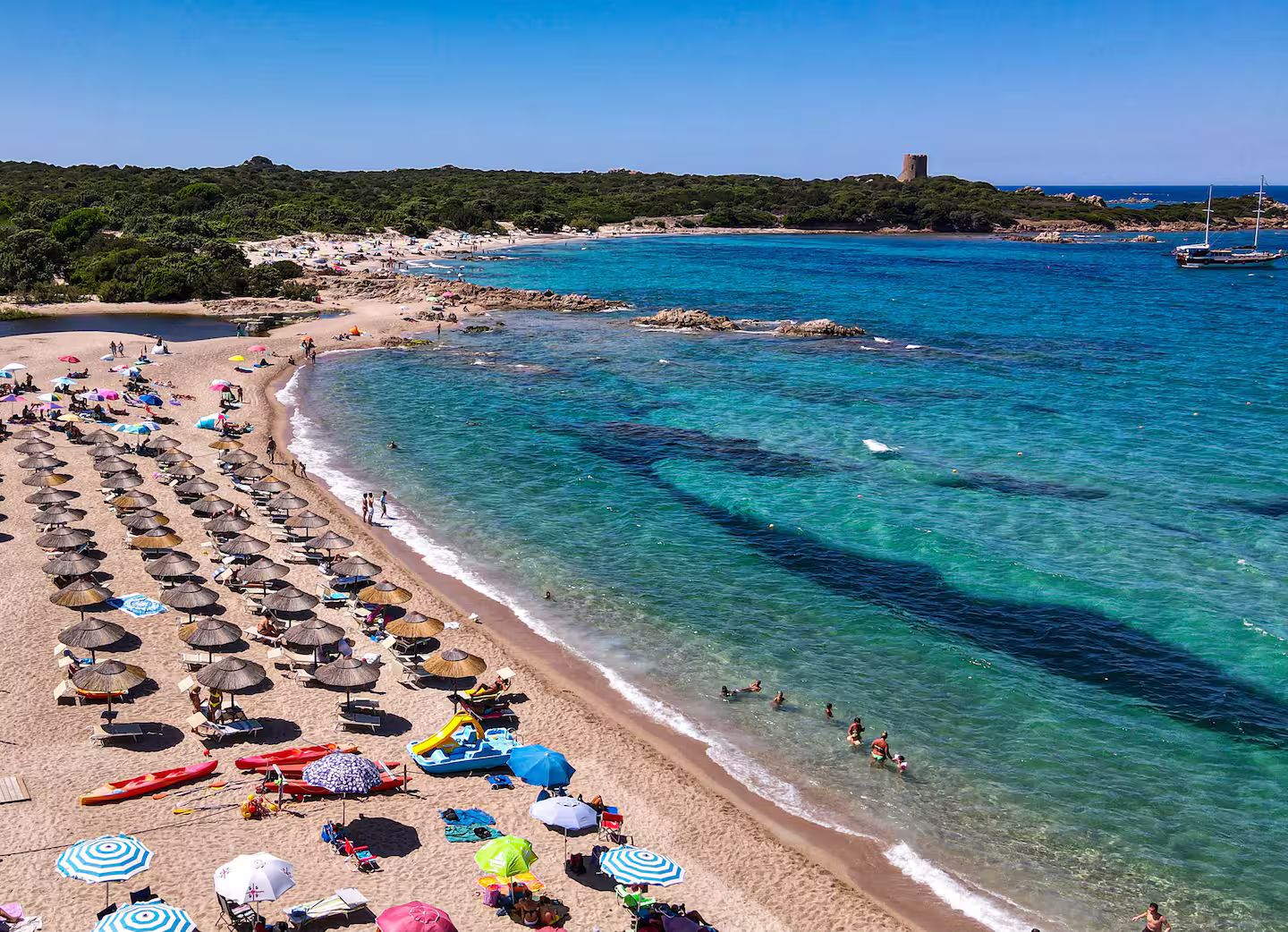 A beach with clear turquoise water, umbrellas, and people.
