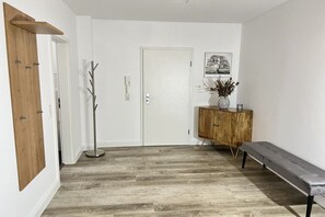 Un corridoio con una porta in legno, una parete bianca, una lampada da terra, un mobile con un vaso e una panca.