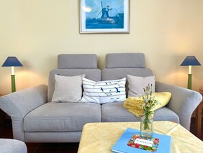 A grey sofa with checkered and striped pillows, a vase with flowers, and a blue book titled 'Gastebuch' on a small table. - Holiday Apartment With 1 Bedroom (Wyk auf Foehr)