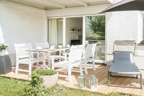 En udendørs terrasse med et bord og stole, en liggestol og en skydedør ind til et indre rum.