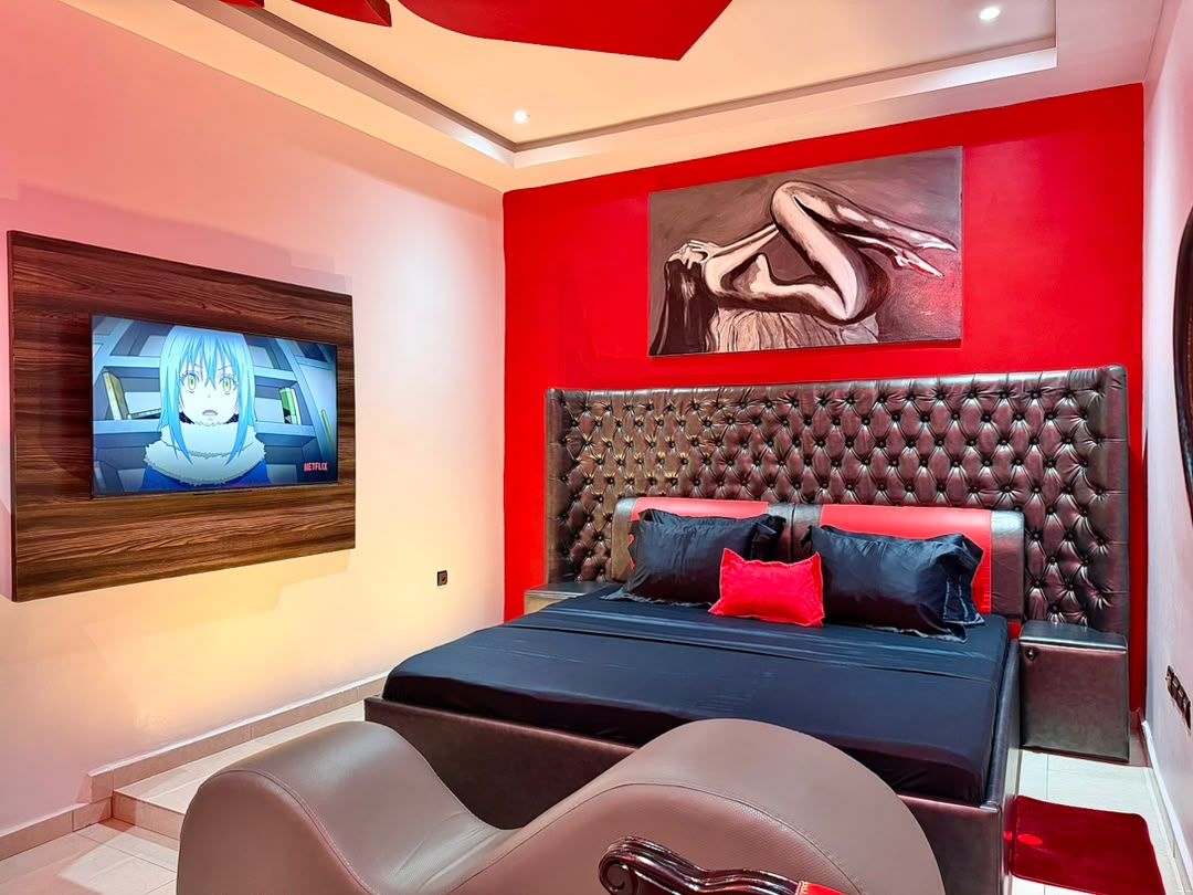 A modern bedroom with a large bed, a mounted TV, and a red accent wall.
