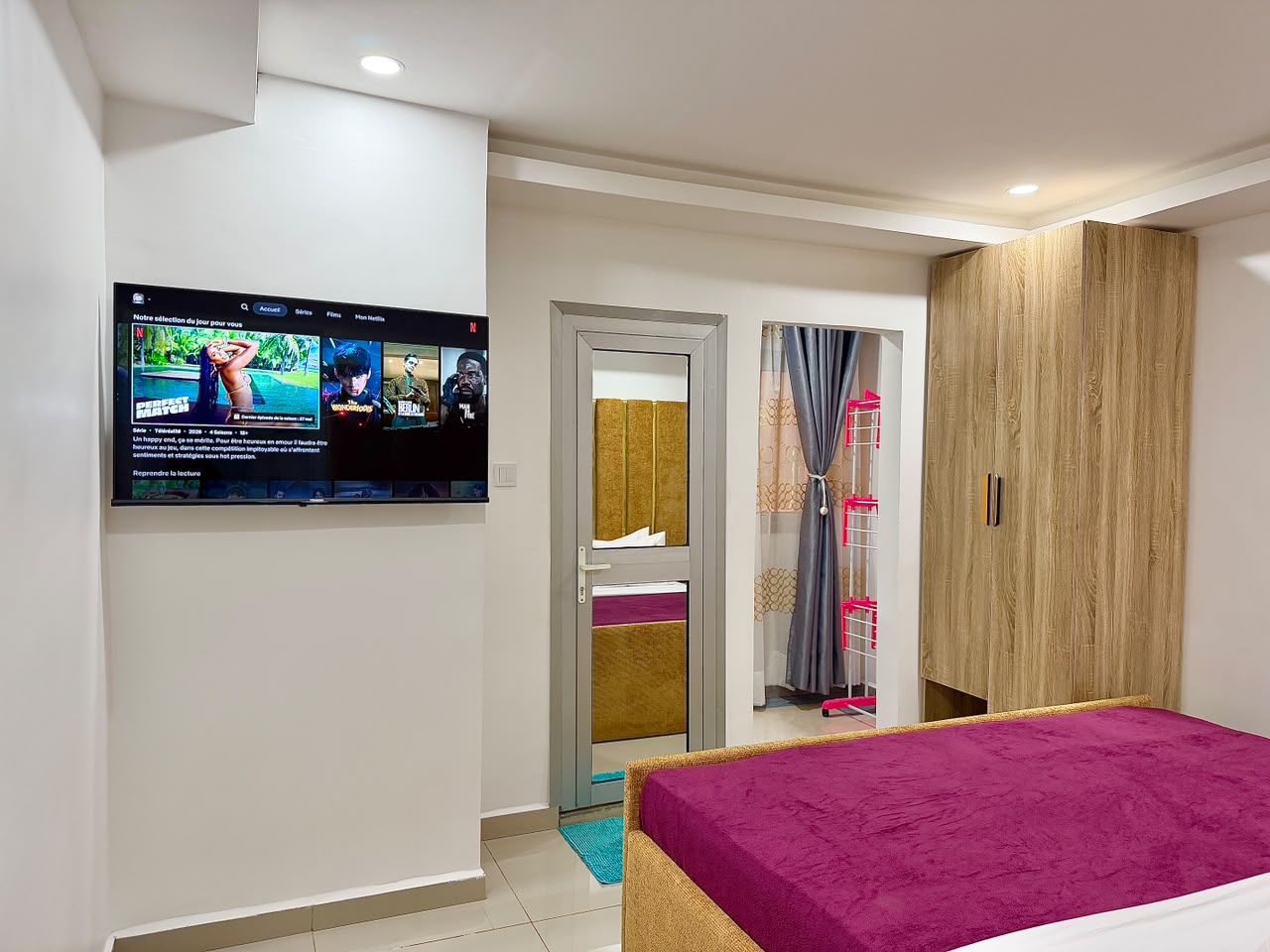 A bedroom with a bed, a TV mounted on the wall, a wardrobe, and a door leading to a bathroom.