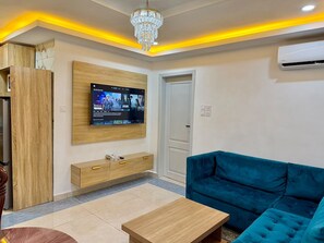 A modern living room with a TV mounted on the wall, a blue sofa, and a wooden cabinet. - Residence la Marina Logpom (Douala)
