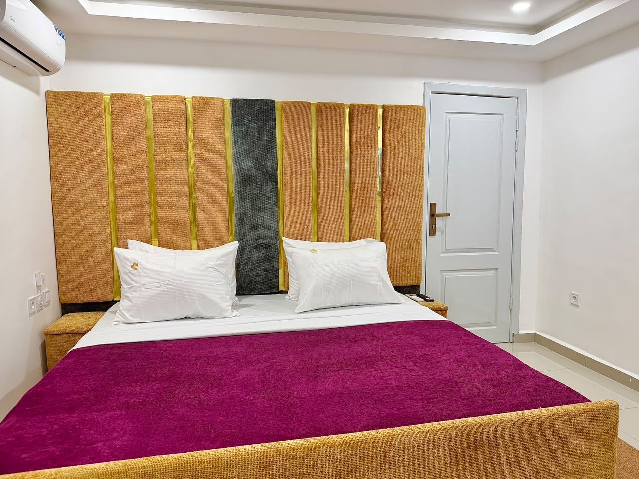 Ein Hotelzimmer mit einem Bett, Kissen, einem Kopfteil mit vertikalen Paneelen, einer Tür und einer weißen Wand.