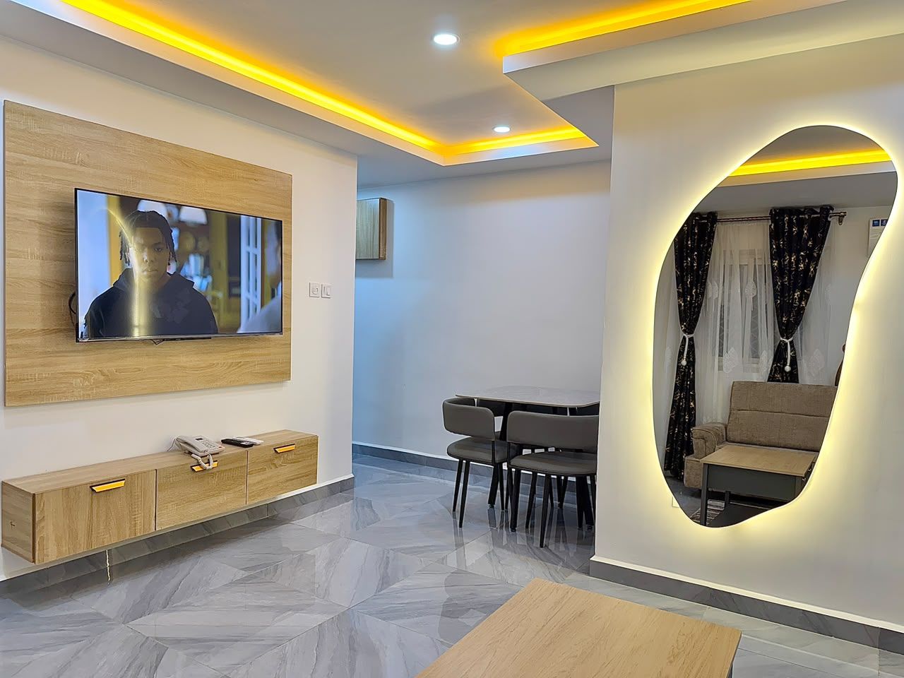Een moderne woonkamer met een flatscreen televisie, een houten tv-meubel, een eethoek met een tafel en stoelen, en een bank.
