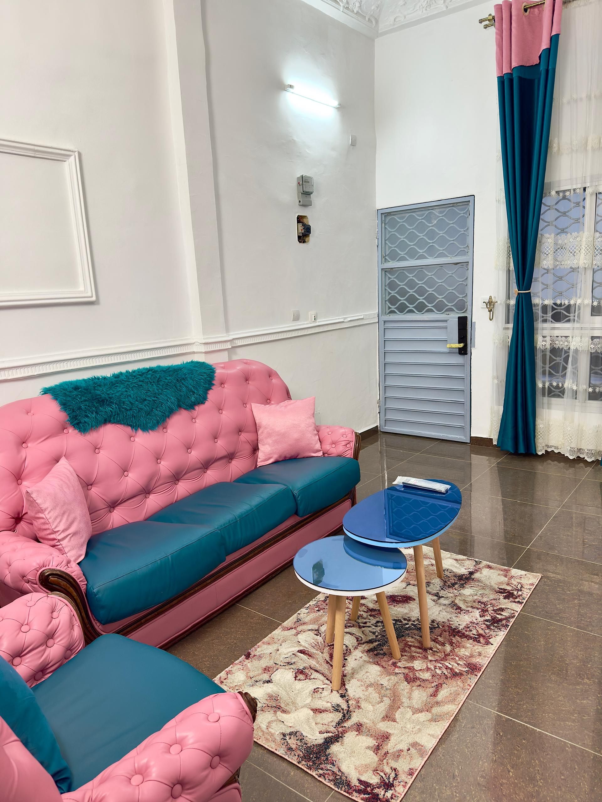 Een kamer met een roze en turquoise bank, een klein blauw tafeltje en een kamerbreed tapijt met een patroon.