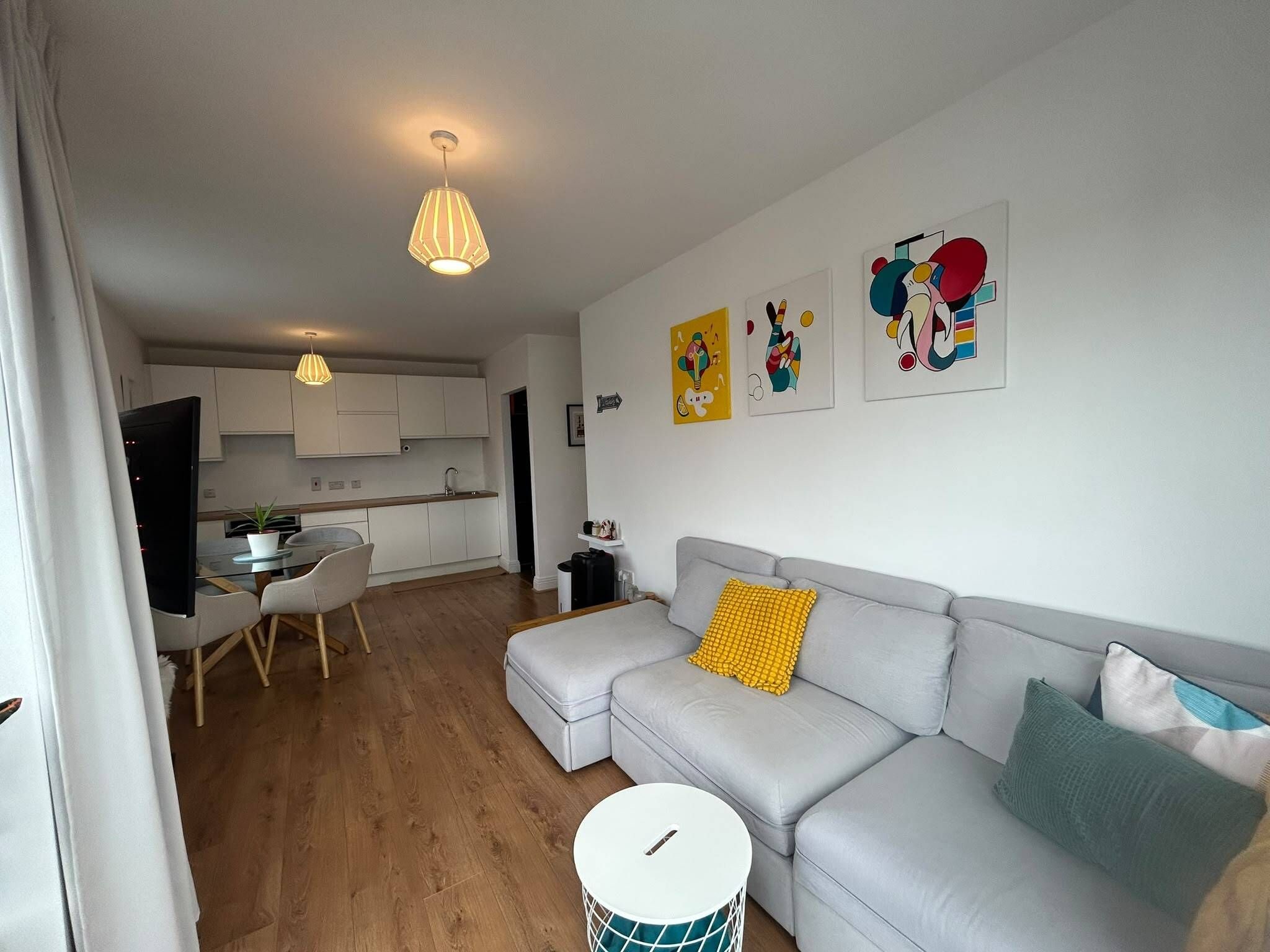 A modern living room with a grey sofa, a dining table with chairs, a kitchen in the background, and a round white table with a drum.