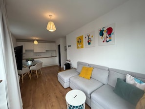 A modern living room with a grey sofa, a dining table with chairs, a kitchen in the background, and a round white table with a drum. - Stylish 2BDR Apt with Free parking in Dublin 8 (Dublin)