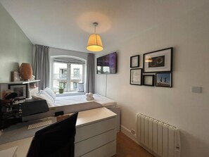 Ett modernt sovrum med en säng, ett skrivbord och en stol, ett fönster med gardiner, ett element och inramade tavlor på väggen.