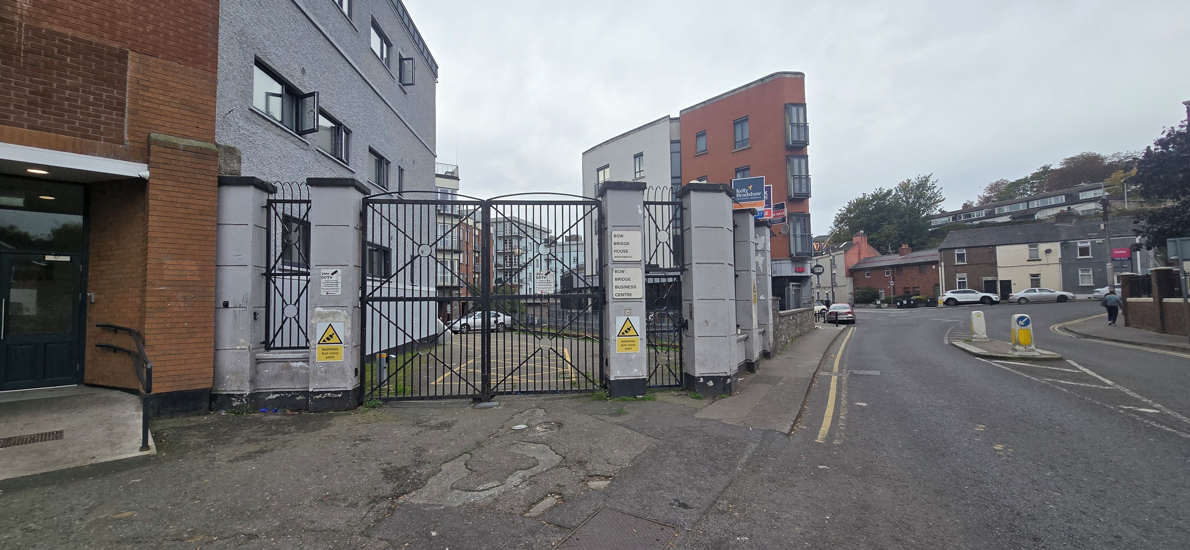A gated entrance to a building with CCTV warning signs, a pedestrian pathway, and parked cars.