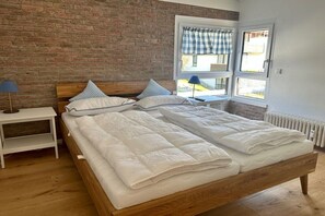 Een tweepersoonsbed met houten frame, een nachtkastje met lamp en een raam met geruit gordijn.