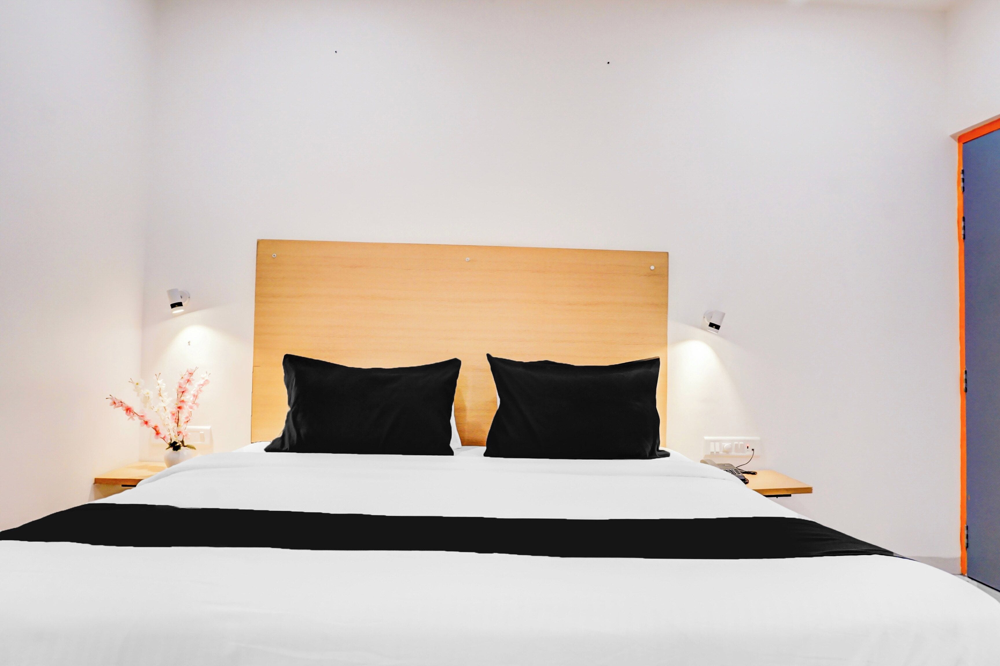Habitación de hotel con cabecera de madera, dos almohadas negras, cubrecama blanco, mesita de noche con lámpara y un jarrón con flores.