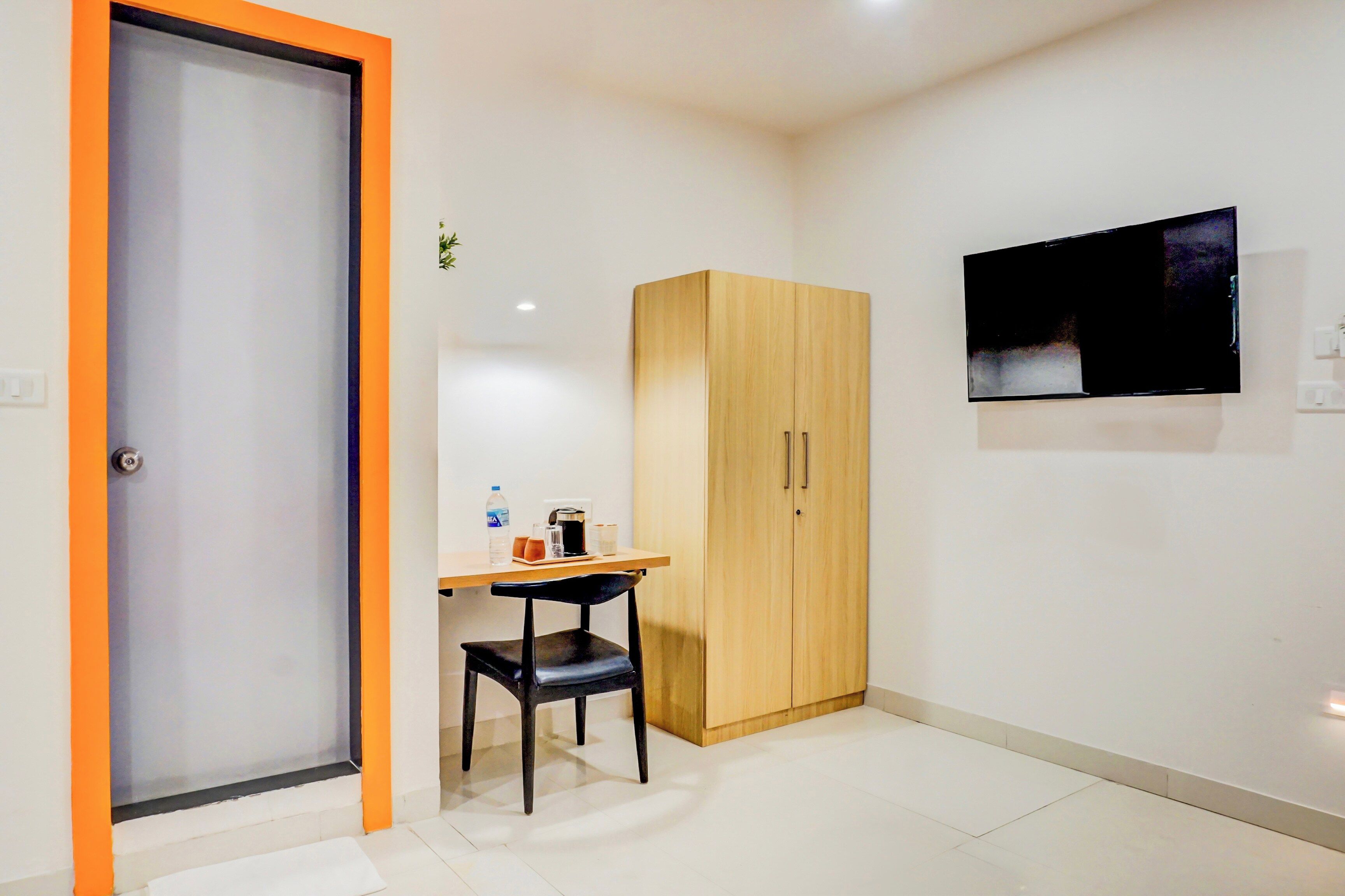 Una habitación pequeña con un armario de madera, un escritorio con una silla, un televisor montado en la pared y una puerta con marco anaranjado.