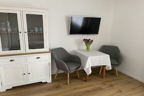 A dining area with a white table, two grey chairs, a cabinet with glass doors, and a flat-screen TV on the wall. - Apartment with 1 bedroom (Berchtesgaden)