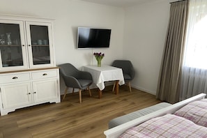 Una habitación con un armario blanco, una mesa con un florero, dos sillas, una cama con un edredón a cuadros y un televisor en la pared.