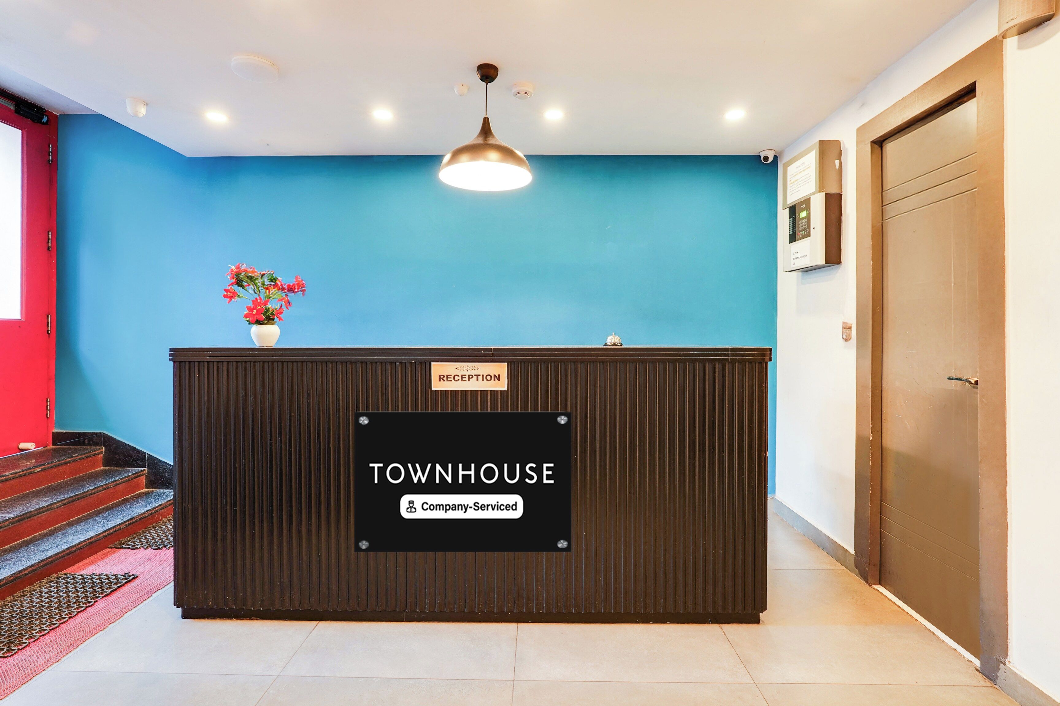 Una recepción con un mostrador identificado como 'TOWNHOUSE', un cartel que dice 'Servicio de la empresa', una pared azul, una puerta de madera y una escalera con barandilla roja.
