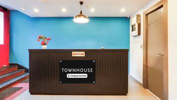 Una recepción con un mostrador identificado como 'TOWNHOUSE', un cartel que dice 'Servicio de la empresa', una pared azul, una puerta de madera y una escalera con barandilla roja.