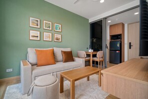 Una sala de estar con un sofá, una mesa de centro y una zona de comedor con sillas. Hay cuadros en la pared, un refrigerador y un microondas.