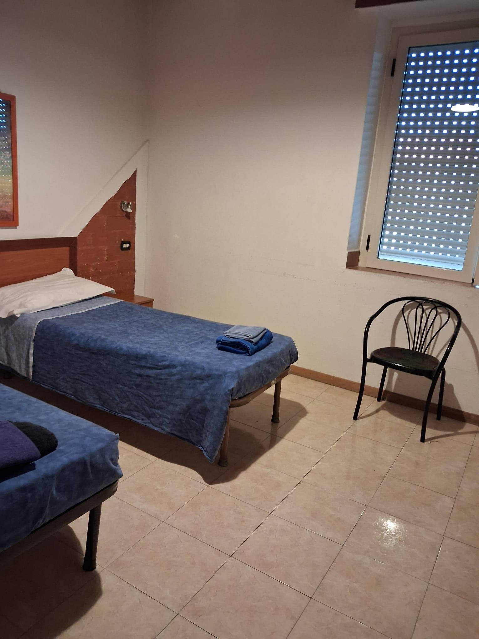 Une chambre avec deux lits simples, une chaise et une fenêtre avec des stores.