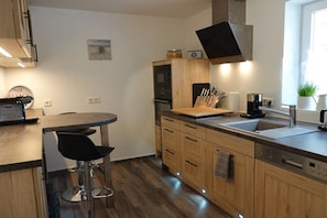A modern kitchen with wooden cabinets, a built-in oven, a coffee machine, and a bar area with stools. - Holiday home with 2 bedrooms (Kappeln Kopperby)