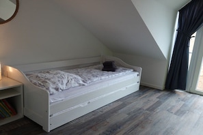 A small, white, single-bed room with a sloped ceiling, a bedside table, and a window with curtains. - Holiday home with 2 bedrooms (Kappeln Kopperby)
