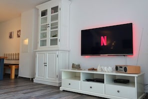 A living room with a flat-screen TV mounted on the wall, a white cabinet with glass doors, and a wooden entertainment unit with electronic devices. - Holiday home with 2 bedrooms (Kappeln Kopperby)