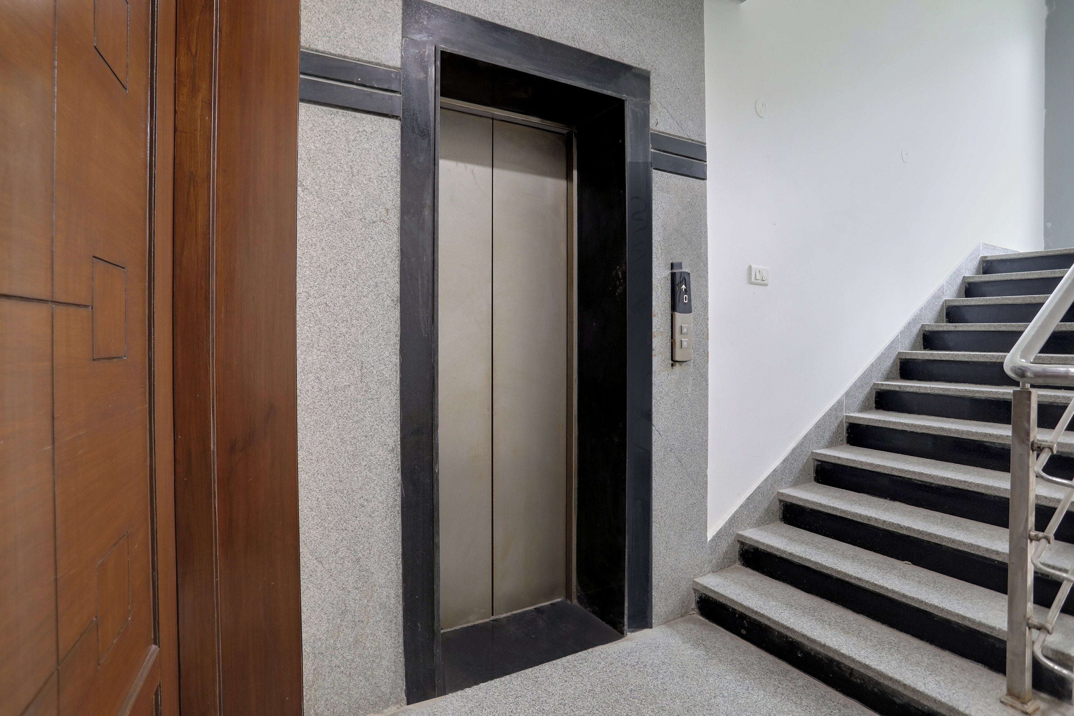 Una puerta de madera con un picaporte vertical, una puerta de ascensor con marco negro y una escalera con barandales metálicos.