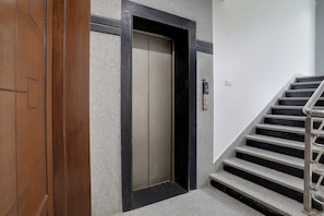 Una puerta de madera con un picaporte vertical, una puerta de ascensor con marco negro y una escalera con barandales metálicos.