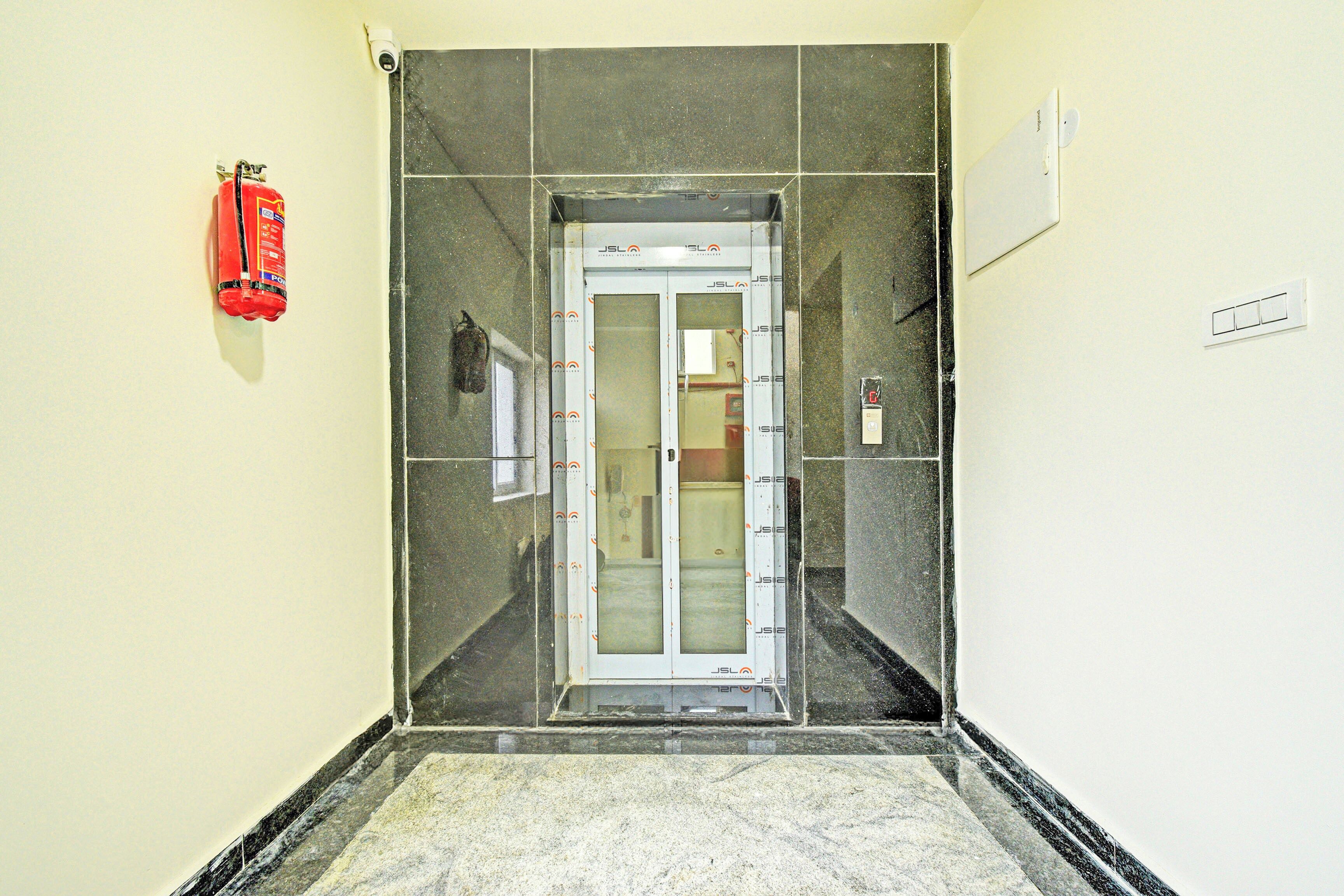 En smal gang med en glaselevatordør, en brandslukker ophængt på væggen og en lyskontakt til højre.