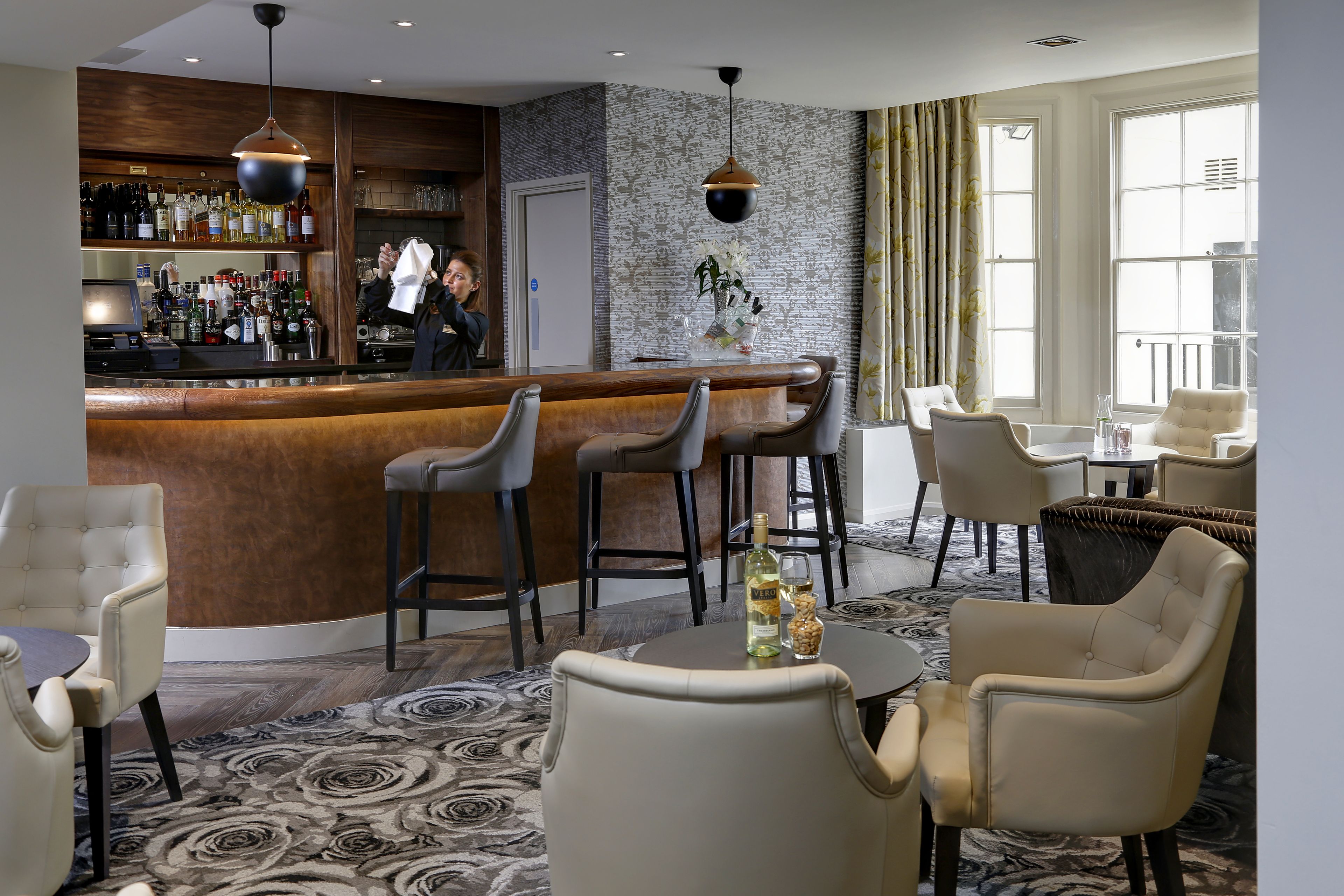Uma área de bar com balcão, banquetas e variedade de garrafas. Cadeiras e uma mesa com garrafa e copos.