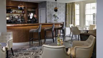 Uma área de bar com balcão, banquetas e variedade de garrafas. Cadeiras e uma mesa com garrafa e copos.