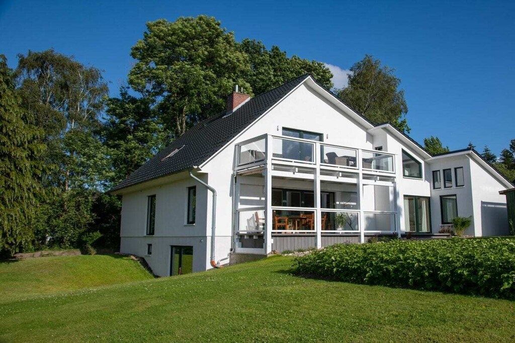 A modern two-story house with a large balcony, surrounded by lush greenery.