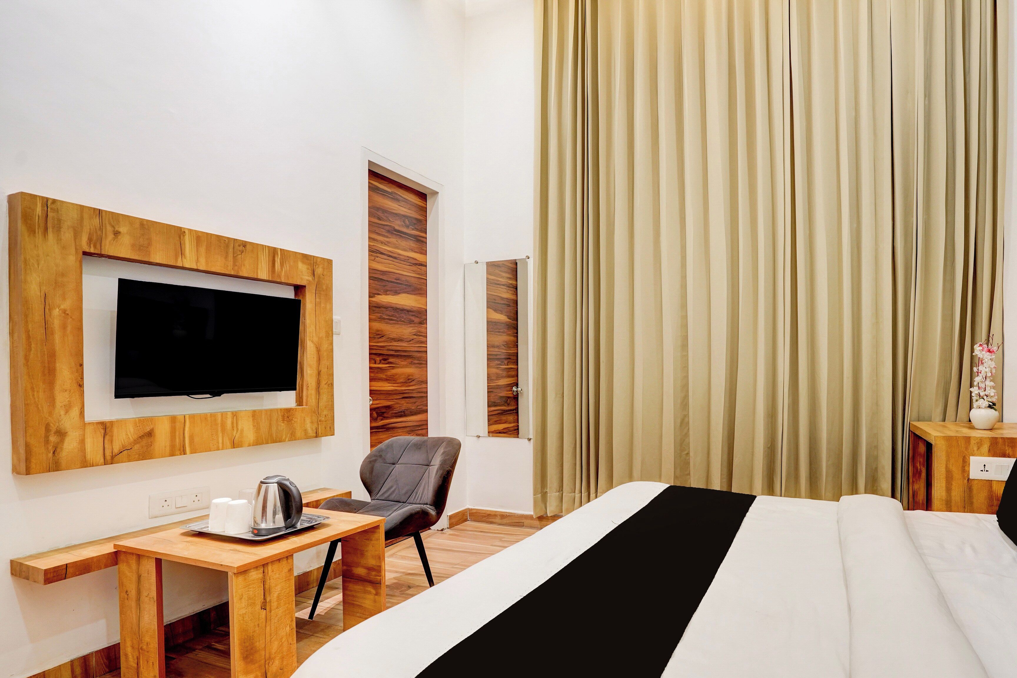 Une chambre d’hôtel avec un lit, un téléviseur fixé sur un panneau en bois, un bureau avec une bouilloire, une chaise et une fenêtre avec des rideaux.