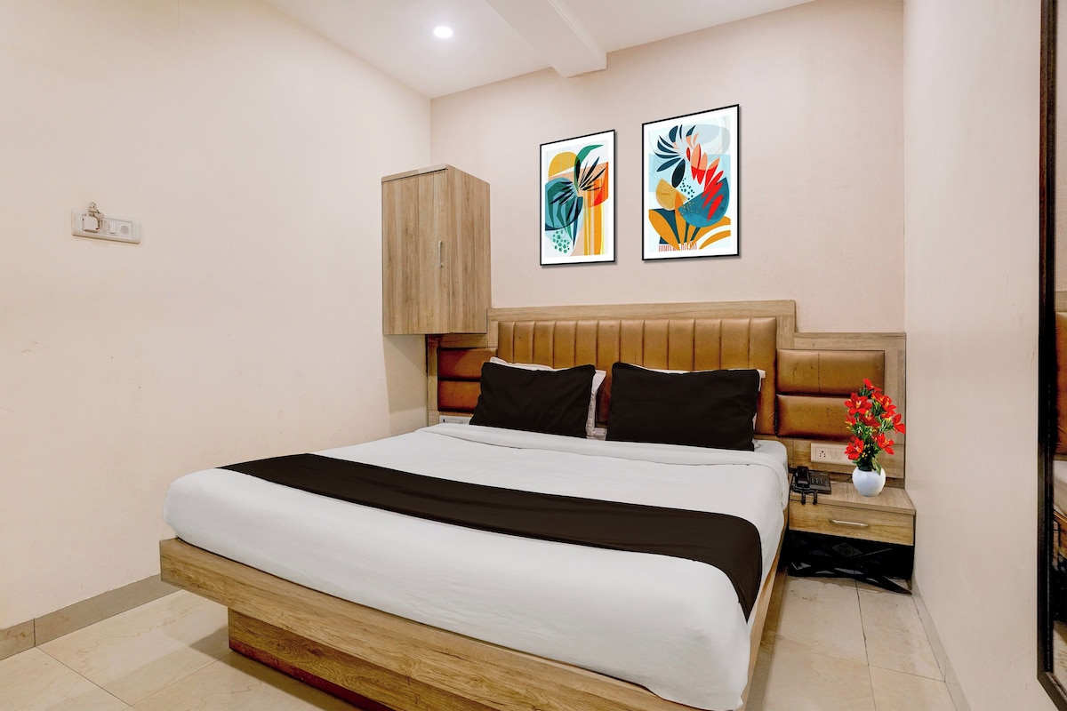 Quarto de hotel com uma cama grande, cabeceira de madeira, duas pinturas abstratas, mesa de cabeceira com um vaso de flores e interruptor de luz na parede.