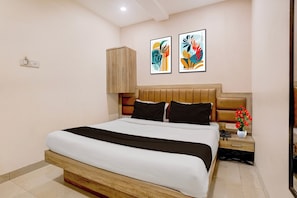 Quarto de hotel com uma cama grande, cabeceira de madeira, duas pinturas abstratas, mesa de cabeceira com um vaso de flores e interruptor de luz na parede.