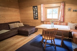 A cozy cabin interior with a wooden floor, a sofa, a dining table with chairs, and a window with curtains. - Holiday home in Rangersdorf with barbecue (Rangersdorf)
