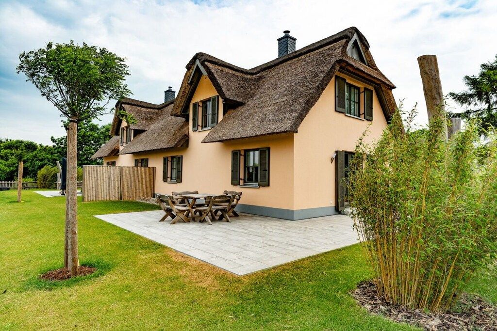 Une maison à deux étages avec un toit de chaume, une terrasse dallée et une table et des chaises en bois.