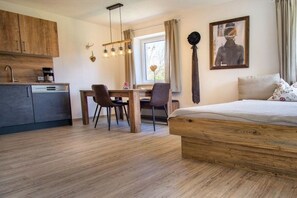 Ein Schlafzimmer mit einem hölzernen Bett, einem Esstisch mit Stühlen, einer Küchenzeile mit Schränken und einem Fenster mit Vorhängen.