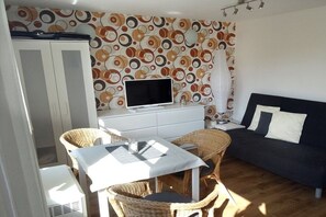 A room with a television on a white cabinet, a dark sofa, wicker chairs, and a patterned wall. - Baltic Sea Idyll Bungalow 4 Seal (Ostseebad Nienhagen)