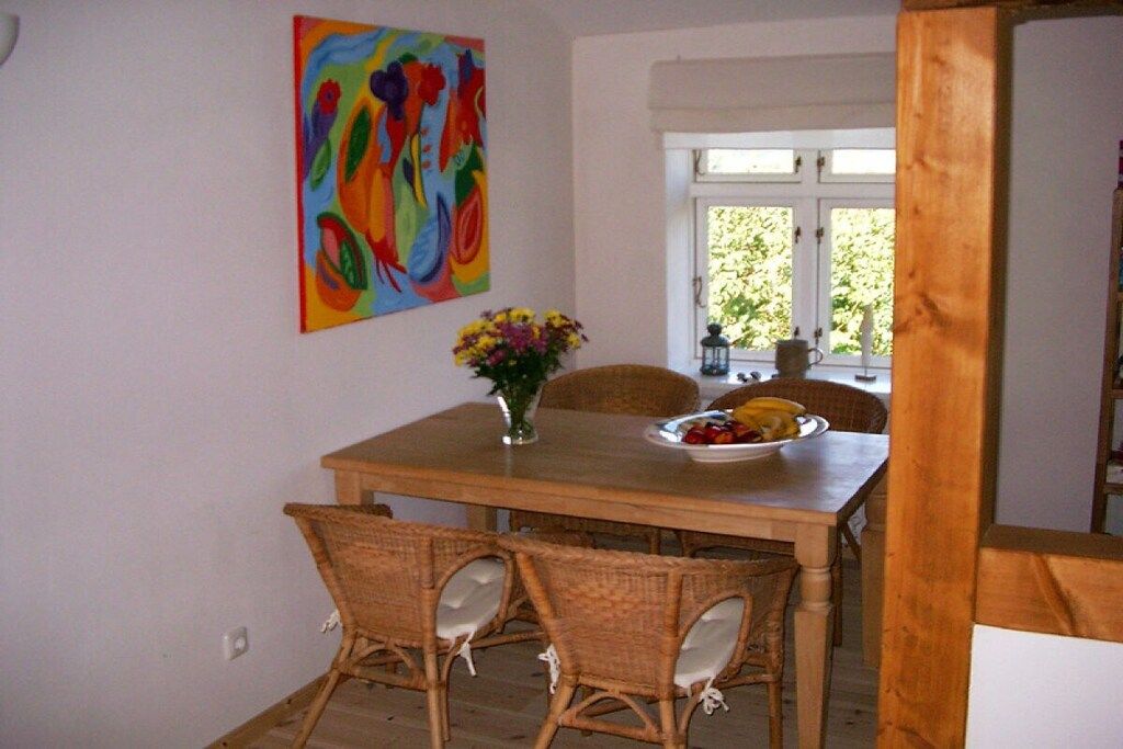 A dining area with a wooden table, wicker chairs, a vase with flowers, and a colorful abstract painting on the wall.