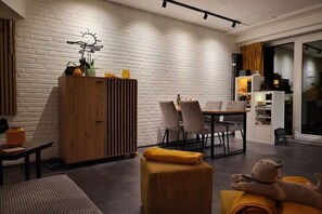 A modern living space with a white brick wall, a wooden cabinet, a dining table with chairs, and a plush sofa with a cat-shaped pillow. - Holiday apartment with 2 bedrooms (Heiligenhafen)