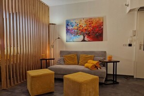 Ein Zimmer mit einem grauen Sofa, zwei gelben Hocker, einem Baumgemälde und einer Holzwand.