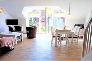 A modern living room with a sofa, a coffee table, a dining table with chairs, a TV, and a large window with a view of buildings outside. - Holiday apartment with 3 bedrooms (Schönhagen)