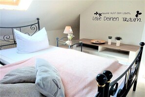 Ein ordentlich bezogenes Bett mit einem weißen Kopfkissen, einer pinkfarbenen Decke und einem grauen Kopfkissen mit Muster. Ein Nachttisch mit Lampe, einer kleinen Pflanze und einer dekorativen Platte sind zu sehen. Eine Wand mit einem motivierenden Zitat ist sichtbar.