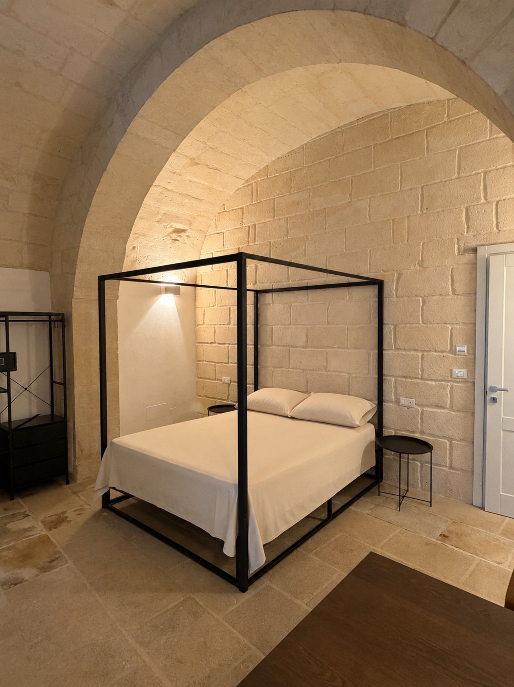 Ein Bett mit Baldachin, ausgestattet mit weißer Bettdecke und Kissen, ein kleiner Nachttisch sowie eine Wandlampe.