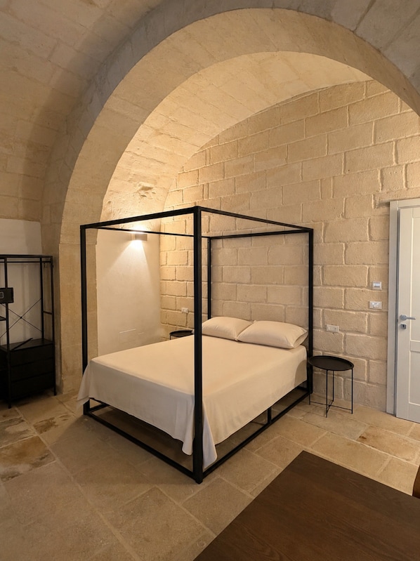 Ein Bett mit Baldachin, ausgestattet mit weißer Bettdecke und Kissen, ein kleiner Nachttisch sowie eine Wandlampe.